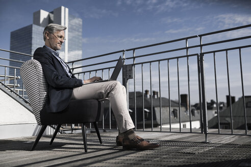 Grauhaariger Geschäftsmann sitzt auf dem Balkon und benutzt einen Laptop - SBOF00733