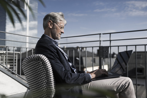 Grauhaariger Geschäftsmann sitzt auf dem Balkon und benutzt einen Laptop, lizenzfreies Stockfoto