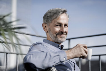 Portrait of smiling grey-haired man sitting on balcony and drinking coffee - SBOF00750