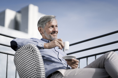 Lächelnder grauhaariger Mann sitzt auf dem Balkon und trinkt Kaffee, lizenzfreies Stockfoto