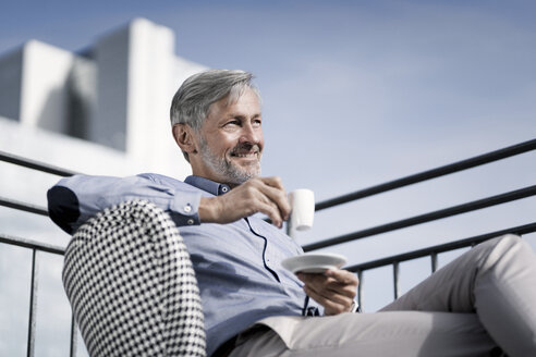 Smiling grey-haired man sitting on balcony and drinking coffee - SBOF00751