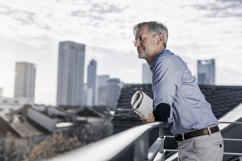 Grauhaariger Mann steht mit Zeitung auf einem Balkon und schaut über die Stadt - SBOF00754