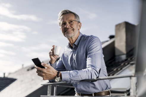Grauhaariger Geschäftsmann mit Smartphone steht auf dem Balkon und trinkt Kaffee - SBOF00755