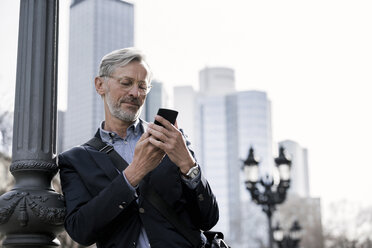 Grey-haired businessman looking at smartphone standing next to street lamp - SBOF00763