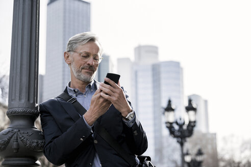 Grauhaariger Geschäftsmann, der neben einer Straßenlaterne steht und auf sein Smartphone schaut - SBOF00763