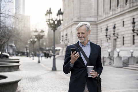 Grauhaariger Geschäftsmann in der Stadt mit Smartphone und Kaffee zum Mitnehmen, lizenzfreies Stockfoto