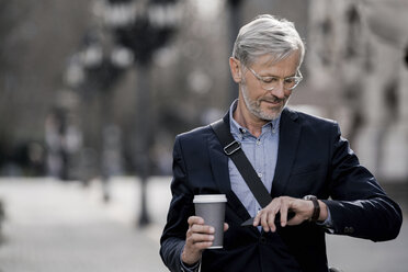 Grey-haired businessman in the city looking at watch holding coffee to go - SBOF00765