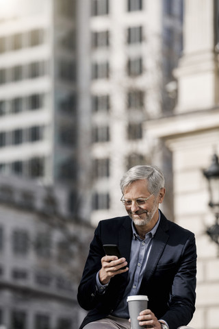 Grauhaariger Geschäftsmann in der Stadt mit Smartphone und Kaffee zum Mitnehmen, lizenzfreies Stockfoto