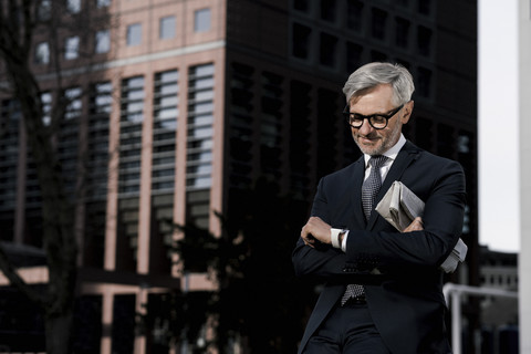 Grauhaariger Geschäftsmann vor rotem Wolkenkratzer, der auf seine Smartwatch schaut, lizenzfreies Stockfoto