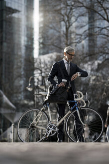 Grauhaariger Geschäftsmann mit Fahrrad und Coffee to go in der Stadt mit Blick auf die Smartwatch - SBOF00787