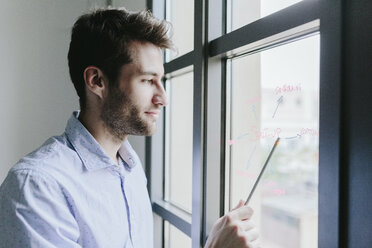 Young businessman standing by office window, looking at organigram - GIOF03273