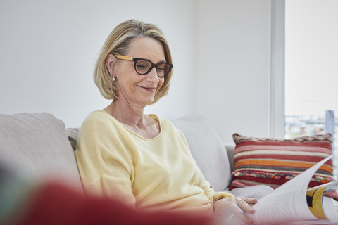 Lächelnde reife Frau zu Hause auf dem Sofa beim Lesen einer Zeitschrift, lizenzfreies Stockfoto