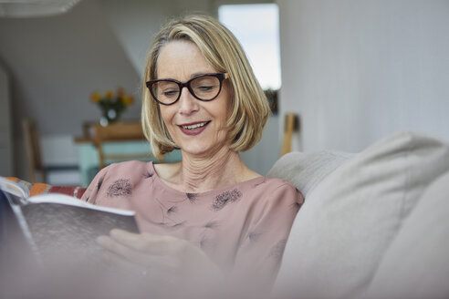 Lächelnde reife Frau zu Hause auf dem Sofa beim Lesen eines Buches - RBF06062