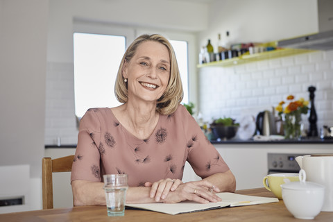 Porträt einer lächelnden reifen Frau mit Notizbuch am Küchentisch, lizenzfreies Stockfoto