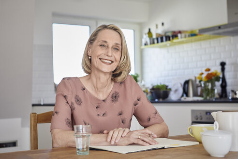 Porträt einer lächelnden reifen Frau mit Notizbuch am Küchentisch - RBF06075