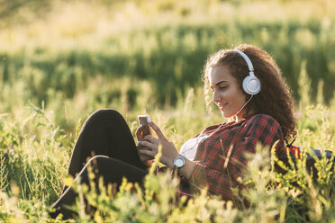 Teenager-Mädchen hört Musik mit Kopfhörern auf einer Wiese und schaut auf ihr Handy - VPIF00116