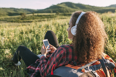 Teenager-Mädchen mit Rucksack hört Musik mit Kopfhörern auf einer Wiese - VPIF00118