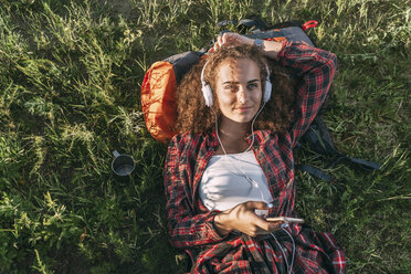 Teenager-Mädchen mit Rucksack auf einer Wiese liegend und mit Kopfhörern und Handy Musik hörend - VPIF00120