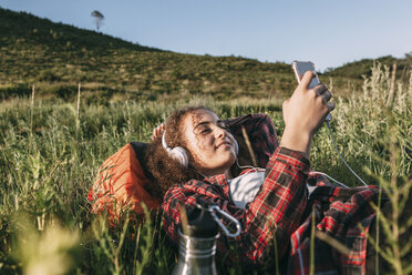 Teenage girl with backpack lying on a meadow listening music with headphones and cell phone - VPIF00121