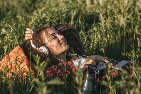 Teenager-Mädchen mit Rucksack auf einer Wiese liegend und mit Kopfhörern und Handy Musik hörend, lizenzfreies Stockfoto