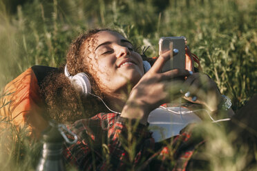 Teenager-Mädchen mit Rucksack auf einer Wiese liegend und mit Kopfhörern und Handy Musik hörend - VPIF00123