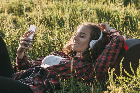 Teenager-Mädchen mit Rucksack auf einer Wiese liegend und mit Kopfhörern und Handy Musik hörend - VPIF00124