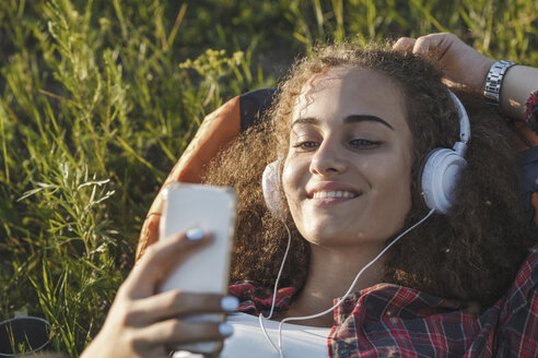 Teenager-Mädchen mit Rucksack auf einer Wiese liegend und mit Kopfhörern und Handy Musik hörend - VPIF00125