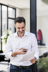 Smiling businessman looking at cell phone and documents - UUF11679