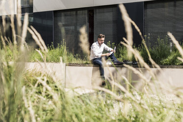 Businessman sitting on wall outside office building reading document - UUF11689