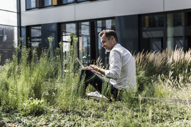 Smiling businessman working in grass outside office building - UUF11690