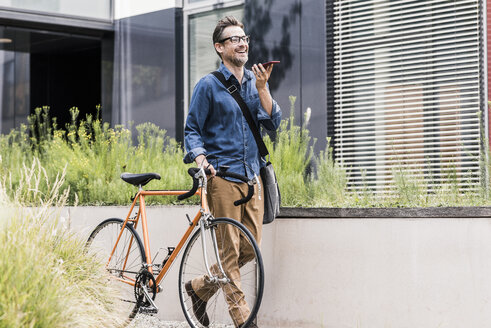 Smiling businessman with cell phone pushing bicycle - UUF11723