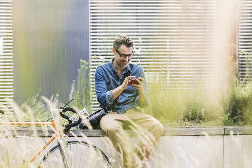 Lächelnder Geschäftsmann, der neben einem Fahrrad ein Mobiltelefon benutzt - UUF11725