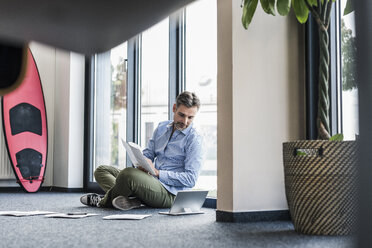 Geschäftsmann arbeitet auf dem Boden im Büro mit Surfbrett lehnt gegen die Wand - UUF11730