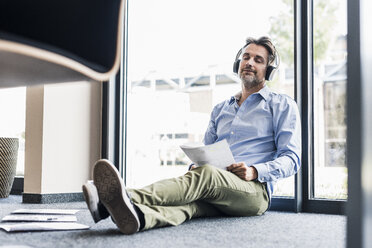 Businessman with headphones sitting on the floor in office - UUF11731