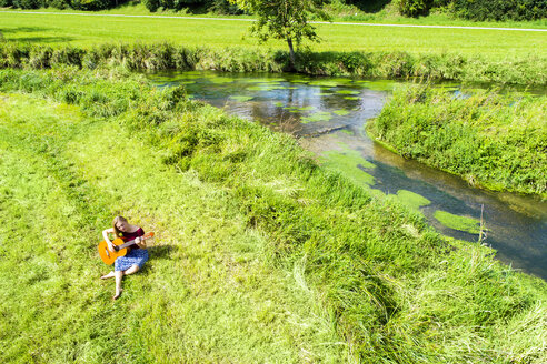 Junge Frau sitzt auf einer Wiese und spielt Gitarre - MAEF12415