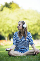 Junge Frau sitzt auf einer Wiese und hört mit Kopfhörern Musik - MAEF12431
