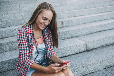 Lächelnde junge Frau sitzt auf einer Treppe und schaut auf ihr Handy - VPIF00158