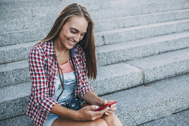 Smiling young woman sitting on stairs looking at cell phone - VPIF00158