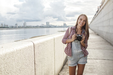 Lächelnde junge Frau mit einer Kamera am Flussufer - VPIF00162