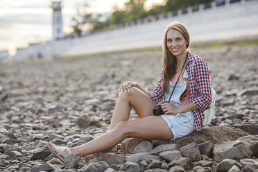 Lächelnde junge Frau mit einer Kamera an einem steinigen Strand - VPIF00164