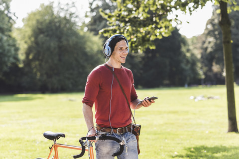 Lachender Mann mit Rennrad, der mit Kopfhörern in einem Park Musik hört, lizenzfreies Stockfoto