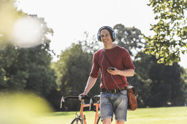 Lächelnder Mann mit Rennrad hört Musik mit Kopfhörern in einem Park - UUF11739