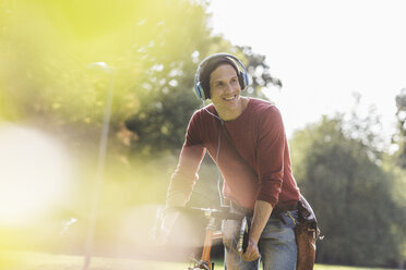 Porträt eines lächelnden Mannes mit Rennrad, der mit Kopfhörern in einem Park Musik hört - UUF11741