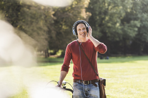 Lachender Mann mit Rennrad, der mit Kopfhörern in einem Park Musik hört, lizenzfreies Stockfoto