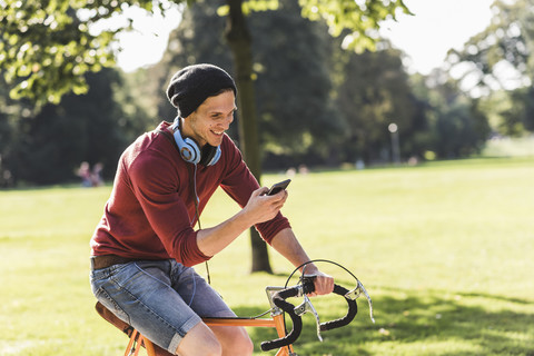 Lachender Mann auf einem Rennrad, der in einem Park auf sein Handy schaut, lizenzfreies Stockfoto