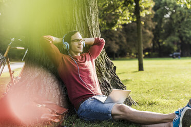 Man with laptop and headphones relaxing in park - UUF11751