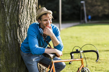 Porträt eines lachenden Mannes mit Rennrad, der sich in einem Park an einen Baumstamm lehnt - UUF11753