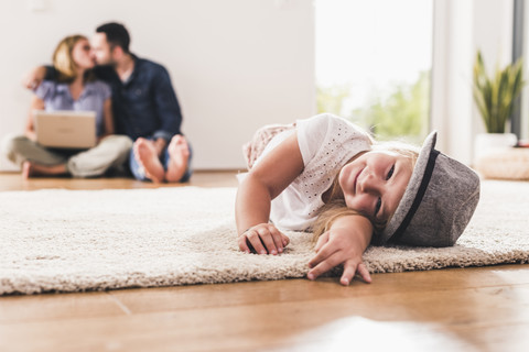 Kleines Mädchen mit Hut, das sich zu Hause amüsiert, Eltern mit Laptop im Hintergrund, lizenzfreies Stockfoto