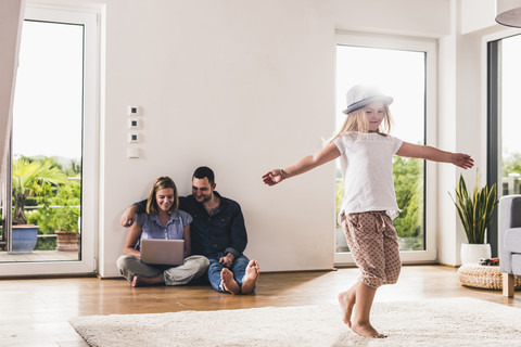 Kleines Mädchen mit Hut, das sich zu Hause amüsiert, Eltern mit Laptop im Hintergrund, lizenzfreies Stockfoto