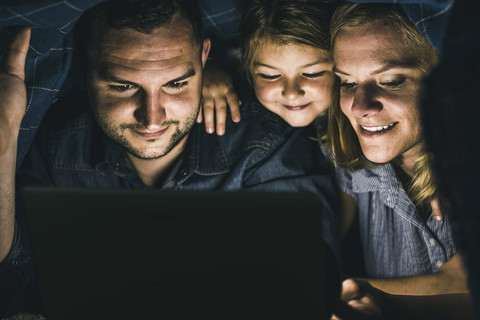 Glückliche Familie, die sich unter einer Decke versteckt und einen Film auf dem Laptop ansieht, lizenzfreies Stockfoto